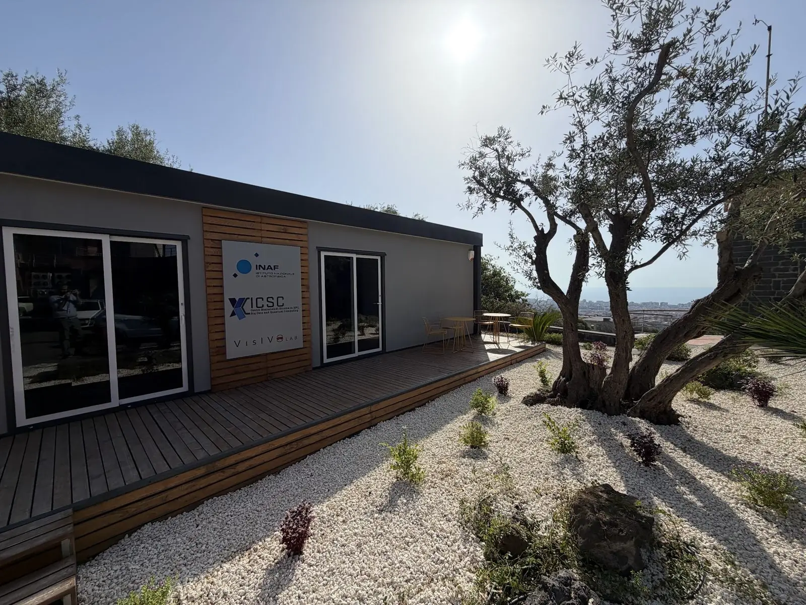 The VisIVO LAB garden: lava stone and white gravel, an olive tree and the pavilion in the sun.